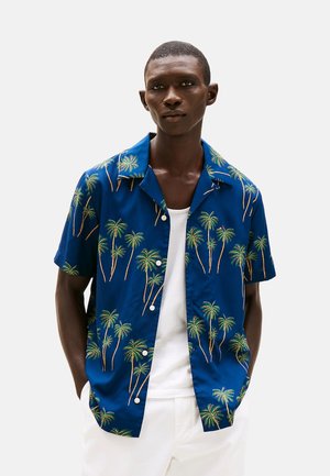 Camisa de manga corta, con botones, de tela azul con un estampado de palmeras en verde y marrón. Textura suave con diseño de cuello.