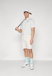 Ανοιχτό γκρι polo μπλούζα με μαύρο λογότυπο Nike, μπεζ σορτς, μέντα πράσινες κάλτσες, λευκό καπέλο και λευκά παπούτσια Nike; κρατώντας μια ράβδο γκολφ.