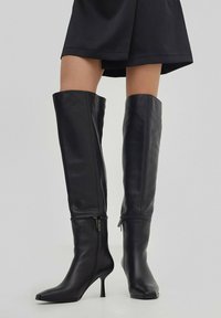 Czarne skórzane botki do kolan z ostrym noskiem i cienkim obcasem typu stiletto. Posiadają zapięcia na zamek przy kostce dla łatwego zakładania.