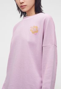 Personne portant une chemise à manches longues texturée violet clair avec un petit motif de fleur d'hibiscus jaune sur le haut gauche de la poitrine.