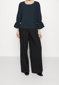 Top blu navy con maniche a sbuffo e scollatura rotonda, abbinato a pantaloni neri a gamba larga e scarpe nere basse. Design semplice e minimale.