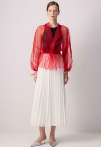 Rote transparente Bluse mit Puffärmeln und gebundenem Bund, kombiniert mit einem weißen Faltenrock und silbernen spitzen Mules.