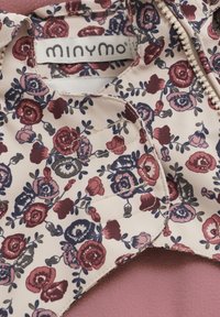 Bloemmotiefstof in tinten rood en blauw op een lichte beige achtergrond, met een ronde kraag en een ritsluiting. Merklabel zichtbaar.