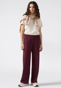 Vrouw met golvend haar, gekleed in een beige t-shirt, een beige trui over haar schouders, bordeauxrode broek en witte sneakers, staande tegen een effen achtergrond.