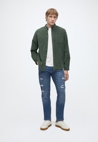 Joven de pie con la mano en el bolsillo, lleva una camisa verde abotonada, camiseta blanca, vaqueros azules rotos y zapatillas blancas sobre un fondo liso.