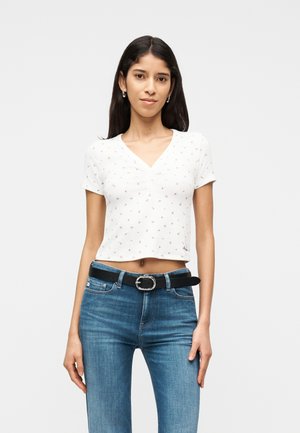 Giovane donna con capelli lunghi e scuri che indossa una maglietta bianca a maniche corte e corta con una piccola stampa floreale e jeans blu con cintura nera.