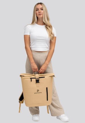 Mochila impermeable beige con cierre enrollable, correas ajustables y un bolsillo frontal con cremallera, que presenta el logo "pactastic" en negro.