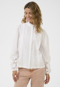 Blouse en coton blanc avec un col haut, des plis verticaux et une bordure en dentelle. Manches longues avec des détails en œillets complexes aux poignets.
