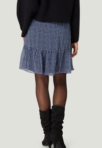 Jupe évasée bleu marine à motif géométrique blanc, volants superposés à l'ourlet, matière transparente, associée à des bottes hautes en daim noir jusqu'au genou.