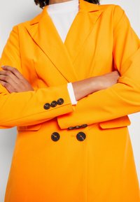 Orange kavaj med skräddarsydd design, slagkragar och svarta knappar på ärmarna. Slät tyg med en ljus, livlig färg.