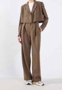 Brązowa krótka marynarka z dwurzędowymi guzikami oraz dopasowane spodnie z wysokim stanem, oba wykonane z gładkiej tkaniny. Pod spodem biała koszulka.