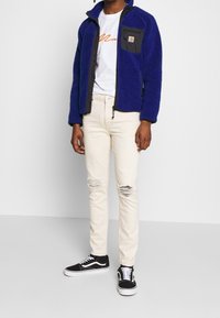 Niebieska kurtka polarowa z czarną kieszenią na zamek, biała koszulka z pomarańczowym napisem, przetarte kremowe dżinsy oraz czarne trampki z białymi detalami.