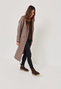 Manteau long en laine marron avec une coupe ajustée, deux poches avant, porté sur un haut sombre et un jean slim, associé à des bottines chelsea marron foncé.