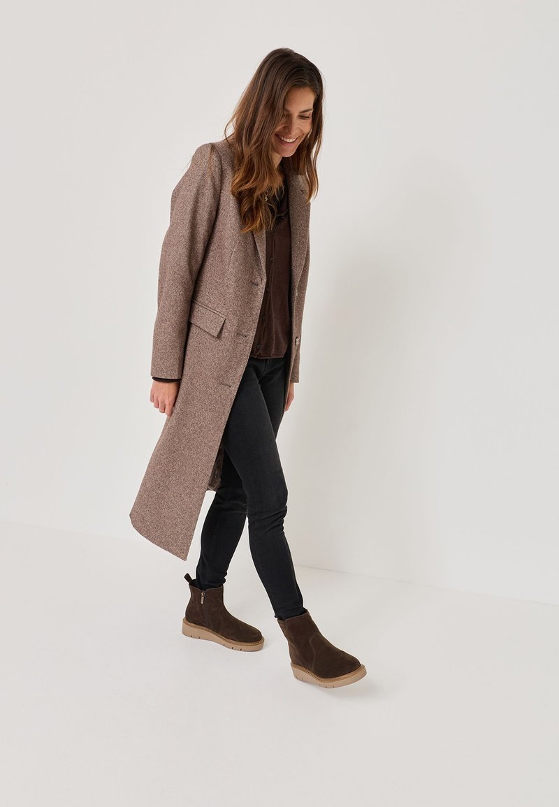 Manteau long en laine marron avec une coupe ajustée, deux poches avant, porté sur un haut sombre et un jean slim, associé à des bottines chelsea marron foncé.