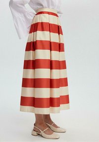 Une jupe plissée longueur cheville avec des rayures alternées rouges et crème, fabriquée en tissu lisse. Associée à des chaussures beige pâle à talons carrés.