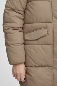 Manteau puffer beige avec texture matelassée, doté d'une poche latérale et d'une coupe décontractée. Les manches sont bouffantes avec des poignets élastiques.