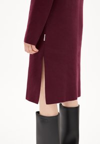 Abito in maglia bordeaux con maniche lunghe, caratterizzato da spacchi laterali. Abbinato a stivali neri in gomma alti fino al ginocchio, contrassegnati da un logo.