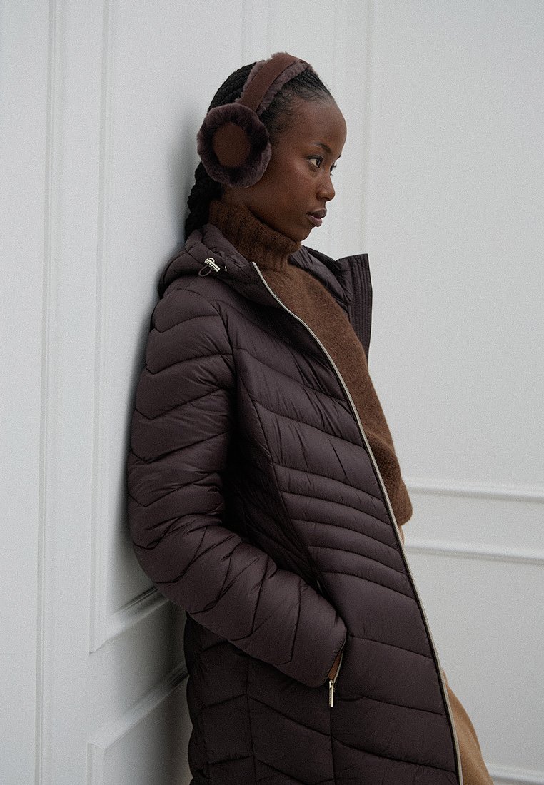 Ciemnobrązowy puchowy płaszcz z przeszyciami w zygzak, zapinany na zamek, z kapturem. Noszony z brązowym swetrem z wysokim kołnierzem i dopasowanymi nausznikami.
