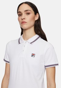 Weißes Polo-Shirt mit einer Knopfleiste aus drei Knöpfen, geripptem Kragen mit roten und marineblauen Streifen sowie einem bestickten Logo auf der linken Brust.