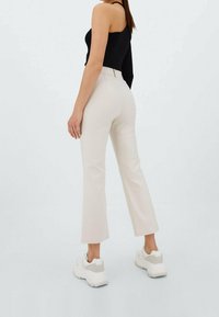 Pantalon évasé beige à texture lisse, taille haute, avec un détail subtil de poche arrière, associé à des baskets blanches et un haut noir.