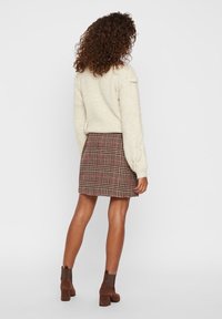 Vrouw met krullend haar in een crème kabelgebreide trui, een geruite minirok en bruine suède enkellaarsjes, staande met haar rug naar de kijker.