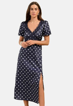Kobieta w granatowej sukience midi w białe groszki, z koronkowym dekoltem w serek, bufkami na rękawach oraz wysokim rozcięciem po boku aż do uda, stojąca z rękami złożonymi.