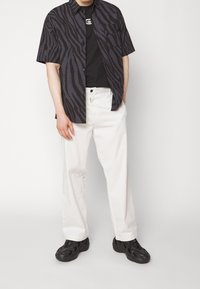 Kortärmad svart skjorta med zebramönster över en svart t-shirt, kombinerad med vita byxor och svarta sneakers, stående mot en neutral bakgrund.