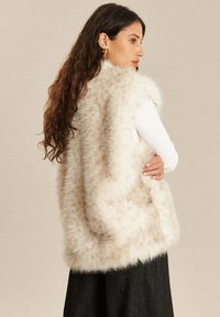 Nepbont vest in roomkleur, met een zachte textuur en een open voorkant. Draagt over een wit longsleeve top, gecombineerd met een donkere rok.