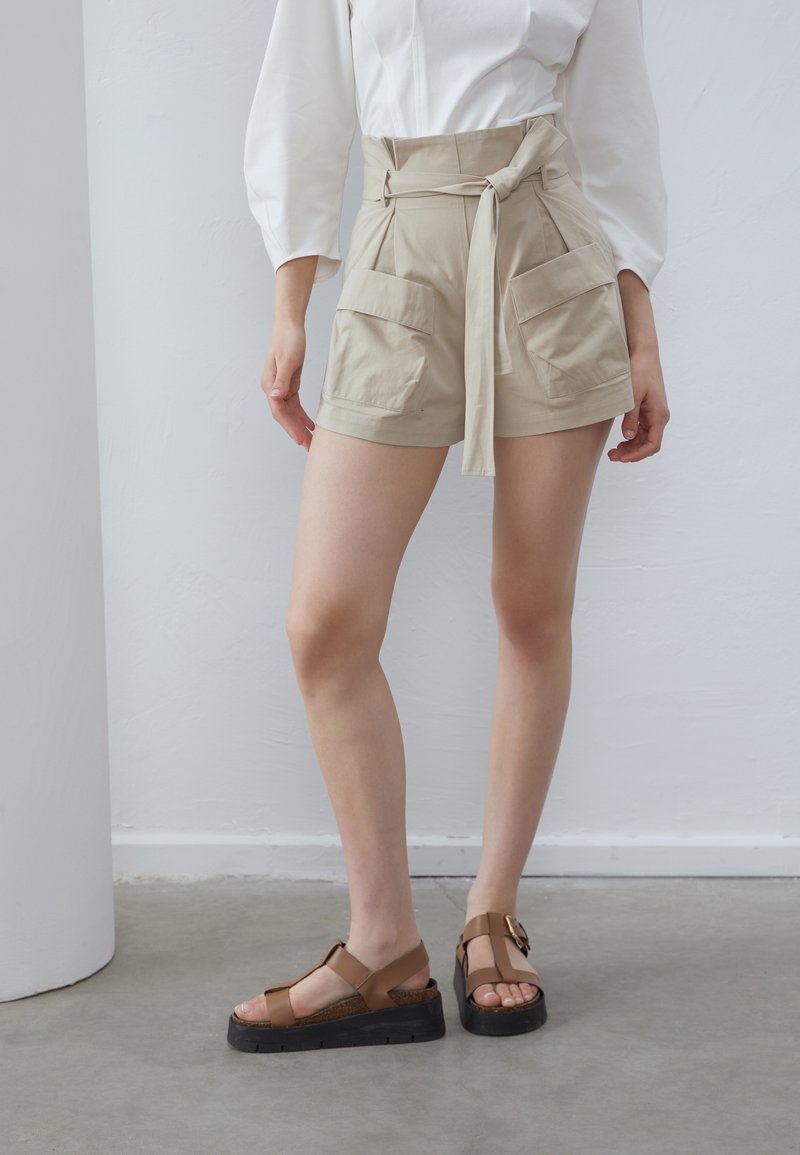 Femme portant un short beige taille haute avec de grandes poches avant et une ceinture nouée, associé à un chemisier blanc et des sandales compensées marron.