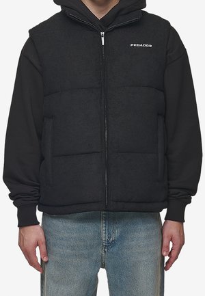 Homme portant une doudoune noire zippée sur un sweat à capuche noir et un jean bleu clair, debout, les bras détendus le long du corps.