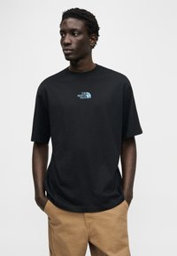 Μαύρο βαμβακερό μπλουζάκι με στρογγυλή λαιμόκοψη, κοντά μανίκια και τουρκουάζ λογότυπο στο στήθος. Το μοντέλο στέκεται σε ουδέτερο φωτισμό, με τα χέρια στις τσέπες.