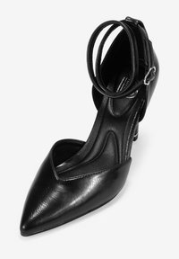 Zapato de tacón alto de cuero negro con punta afilada, correa ajustable en el tobillo y diseño elegante que presenta una textura suave y un detalle mínimo.