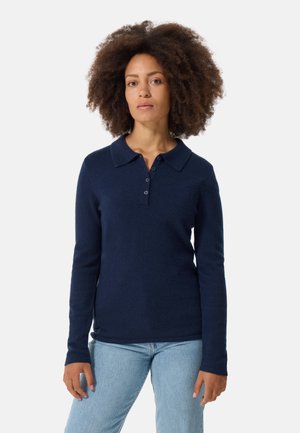 Marineblauwe poloshirt met lange mouwen, gemaakt van zacht materiaal. Voorzien van een kraag, knoopsluiting en een getailleerd ontwerp. Draagt bij lichte blauwe jeans.