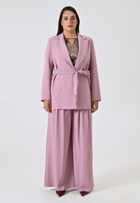 Roze blazer met een strik taille, gecombineerd met wijde roze broeken. Een glanzend bronzen top eronder voegt textuur toe. Gladde stoffenafwerking overal.