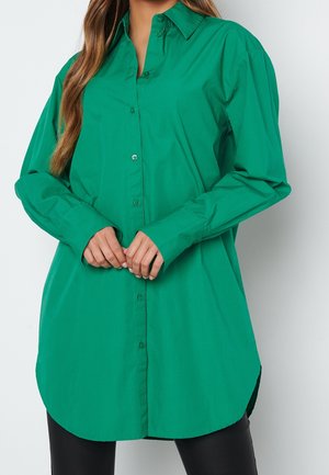 Femme portant une chemise verte ample à manches longues avec col, debout les mains légèrement croisées devant elle.