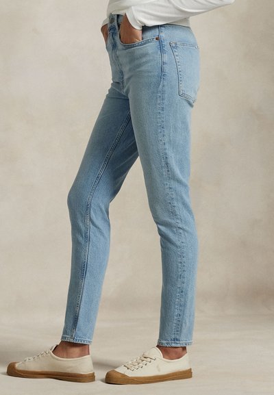Persoon met lichtblauwe slim-fit jeans, witte top met lange mouwen en beige sneakers, staand met handen in de zakken.