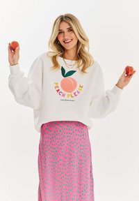 Bílá mikina s nápisem "PEACH PLEASE" a grafikou broskve; v kombinaci s růžovou sukní s motivem zelených teček; drží broskve v obou rukou.