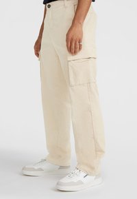 Beige corduroy cargo-housut, joissa on sivutaskut, löysä malli ja suora lahje, yhdistettynä valkoisiin tennareihin, joissa on harmaita yksityiskohtia.