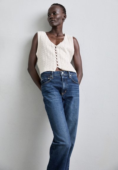 Lichtbeige gebreid mouwloos vest met een V-hals en knoopsluiting, gecombineerd met donkerblauwe rechte jeans. Soepele textuur, casual stijl.