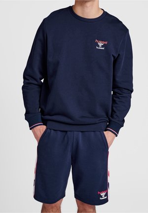 Homme portant un sweat-shirt hummel bleu marine et un short assorti avec des détails en rayures rouges, blanches et bleues, les mains dans les poches, debout devant un fond gris.