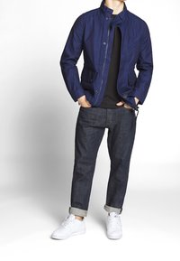 Chaqueta ligera azul marino con cuello alto, múltiples bolsillos delanteros y un diseño ajustado, combinada con pantalones vaqueros oscuros y zapatillas blancas.