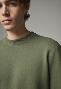 Sweter w oliwkowej zieleni, dziany, z prążkowanym dekoltem; faktura tkaniny; zbliżenie pokazujące szczegóły szwu na ramieniu i dekoltu.