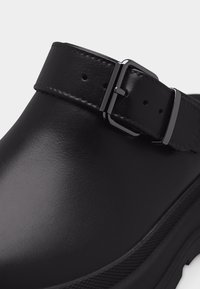 Gros plan sur une sabots en cuir noir avec une lanière cousue et une boucle métallique réglable sur le dessus.