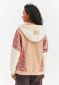 Bluza z kapturem w beżowym i kremowym kolorze o miękkiej teksturze. Posiada różowy panel z białymi kropkami. Etykieta na górnej części pleców dodaje charakterystyczny detal.