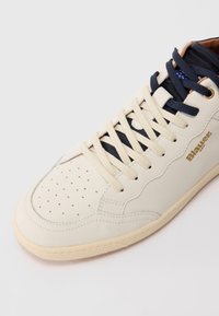 Béžová kožená teniska s námorníckymi modrými šnúrkami, perforovaným dizajnom na špičke a zlatým logom. Gumená podrážka s textúrovanými detailmi.