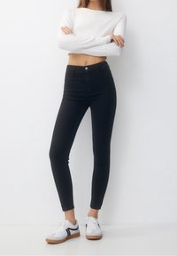 Mujer con los brazos cruzados, vistiendo una camiseta corta de manga larga blanca, jeans skinny negros de cintura alta y zapatillas blancas con suelas de goma.