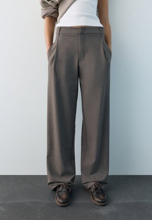 Person mit weiten grauen Hosen, weißem Oberteil, beige gestricktem Ärmel und dunkelbraunen Schnürschuhen, stehend mit den Händen in den Taschen.