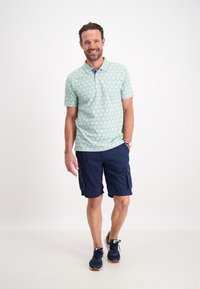 Ljusgrön polo med cirkulärt mönster, korta ärmar och krage; matchas med marinblå cargo shorts. Blå sportskor med vita accenter.