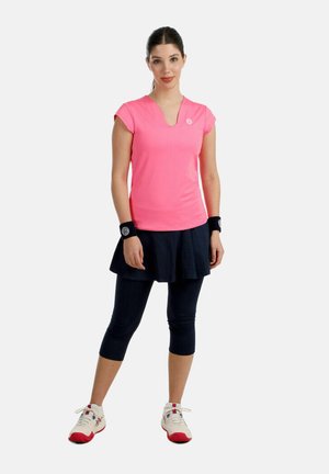 Vrouw staat, draagt een felroze sporttop, een zwarte tennisskirt over leggings, polsbandjes en witte sneakers met rode zolen.