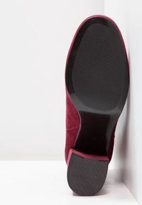 Bordeaux suede schoen met een gestructureerde zwarte rubberen zool, met een gladde, taps toelopende neus en een slanke silhouette, weergegeven vanuit de onderkant.